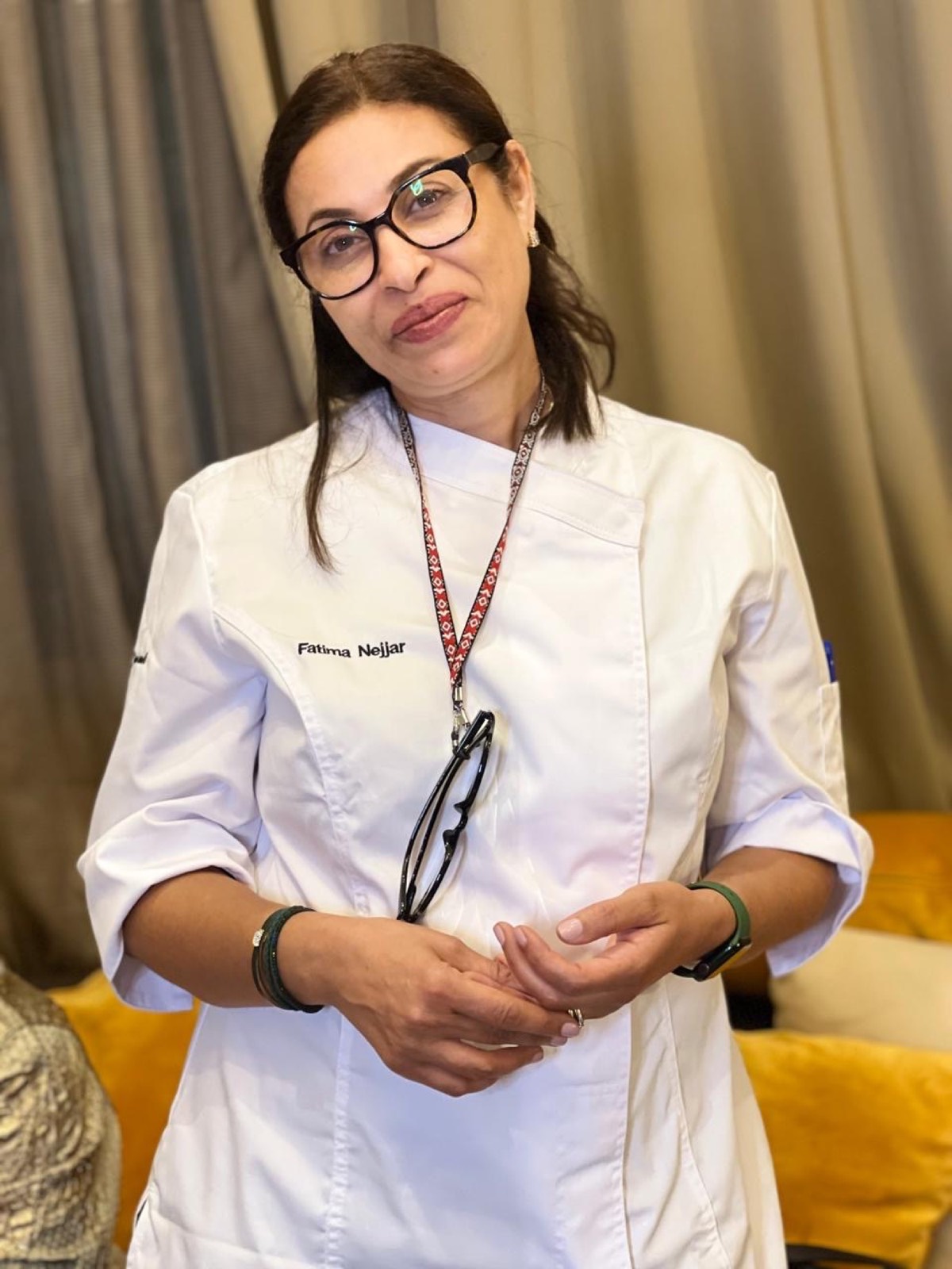 Fatima Nejjar en veste de cuisine blanche brodée à son nom, mains jointes, devant un rideau de lin